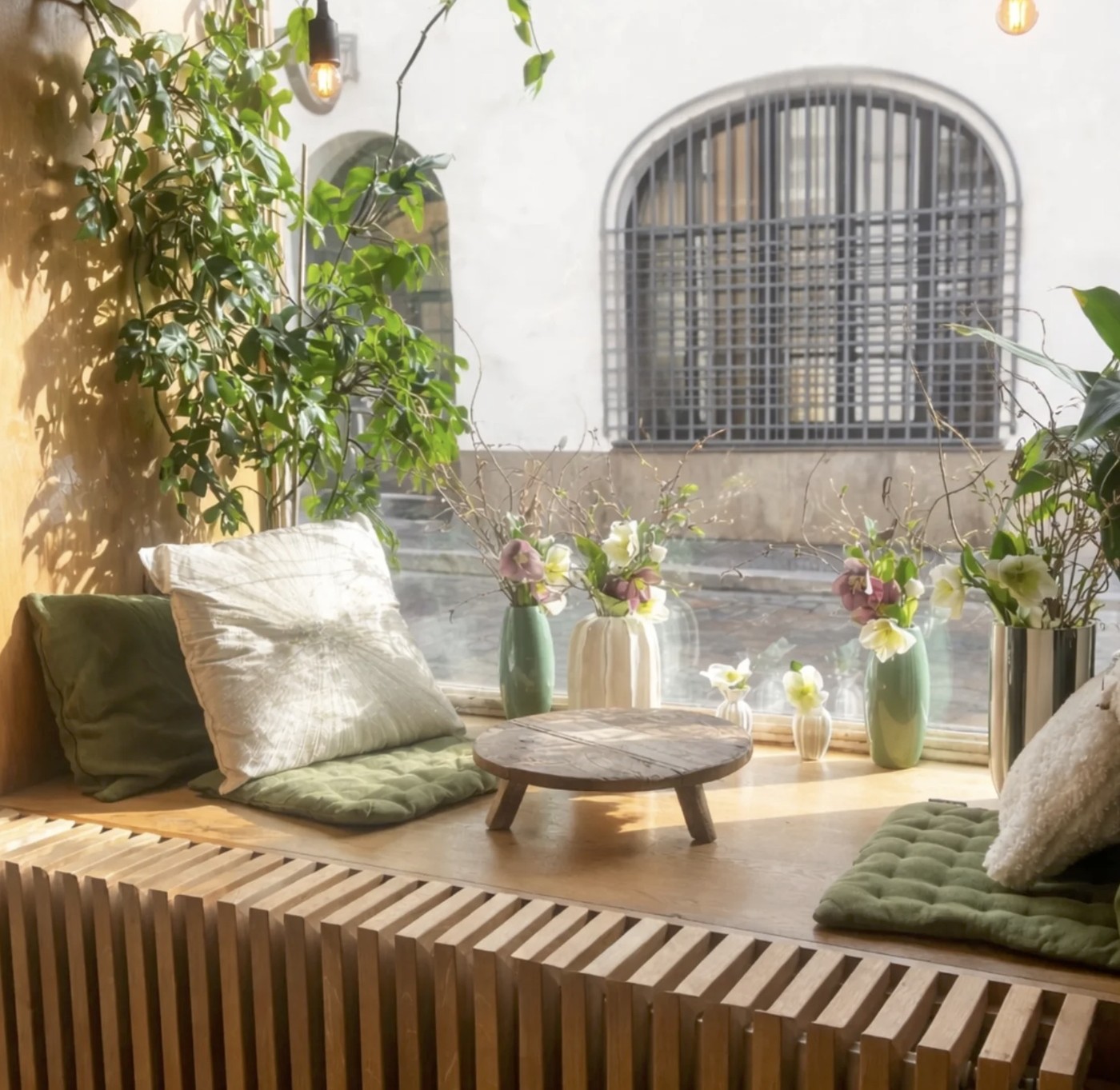 A sunlit window nook at Klobouk dolů with floor cushions, trailing plants, small vases of flowers, and a low wooden table